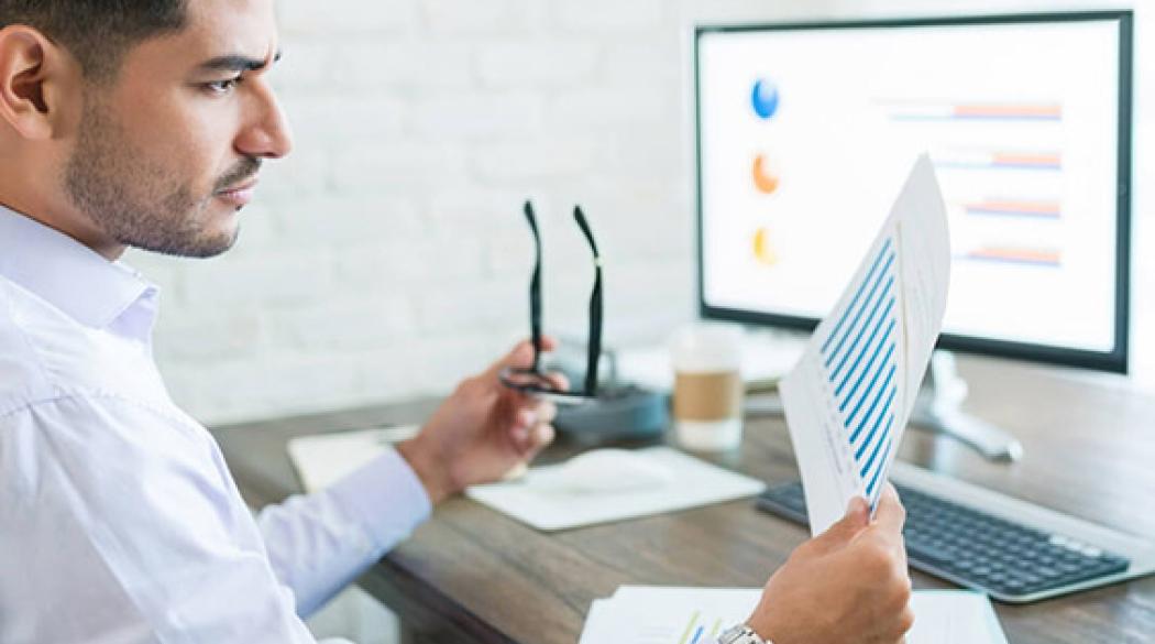 Man holding eyeglasses and paper at computer desk